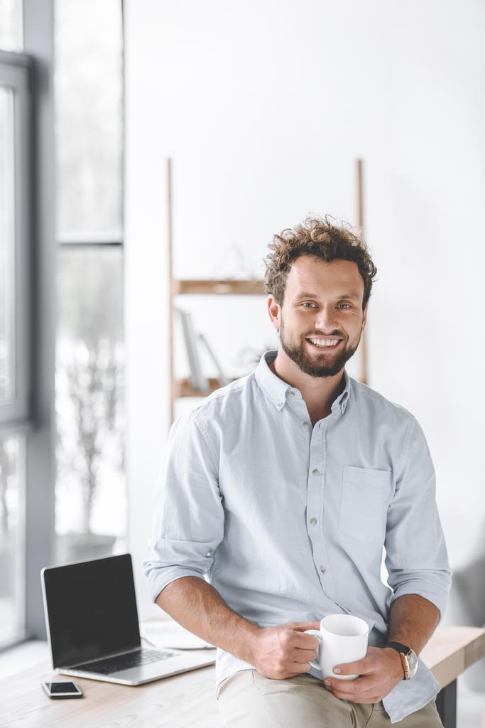 улыбающийся бизнесмен с чашкой кофе сидит на столе с ноутбуком и смартфоном в офисе
 - Фото, изображение