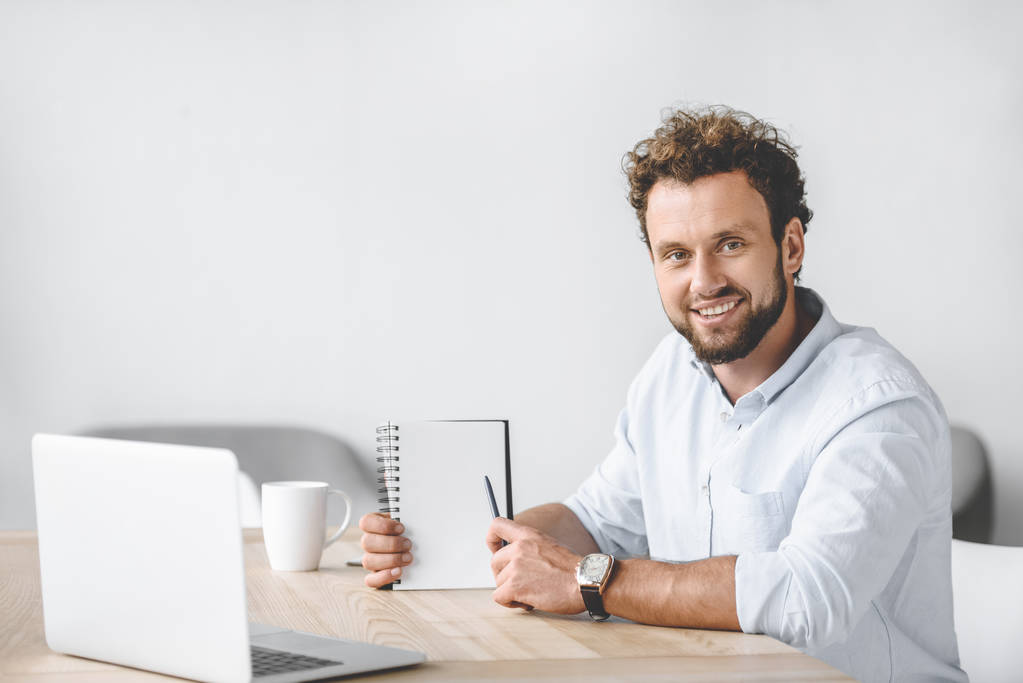 усміхнений бізнесмен сидить на робочому місці з ноутбуком і вказує на порожній блокнот в руці
 - Фото, зображення