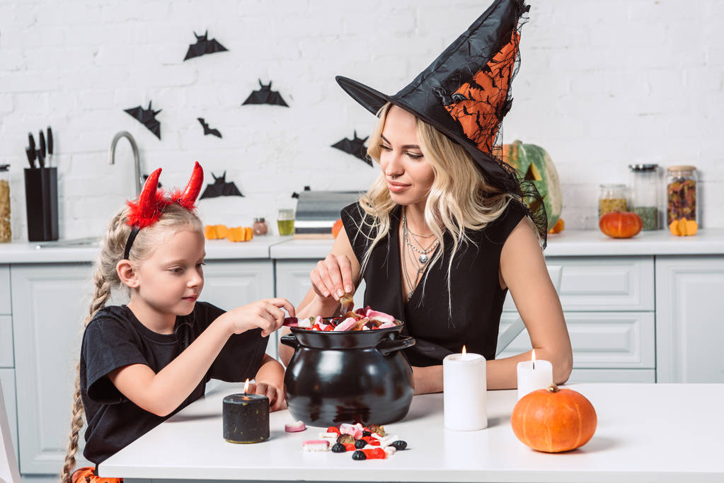 mãe e filhinha em trajes de Halloween à mesa com guloseimas em panela preta na cozinha
 - Foto, Imagem