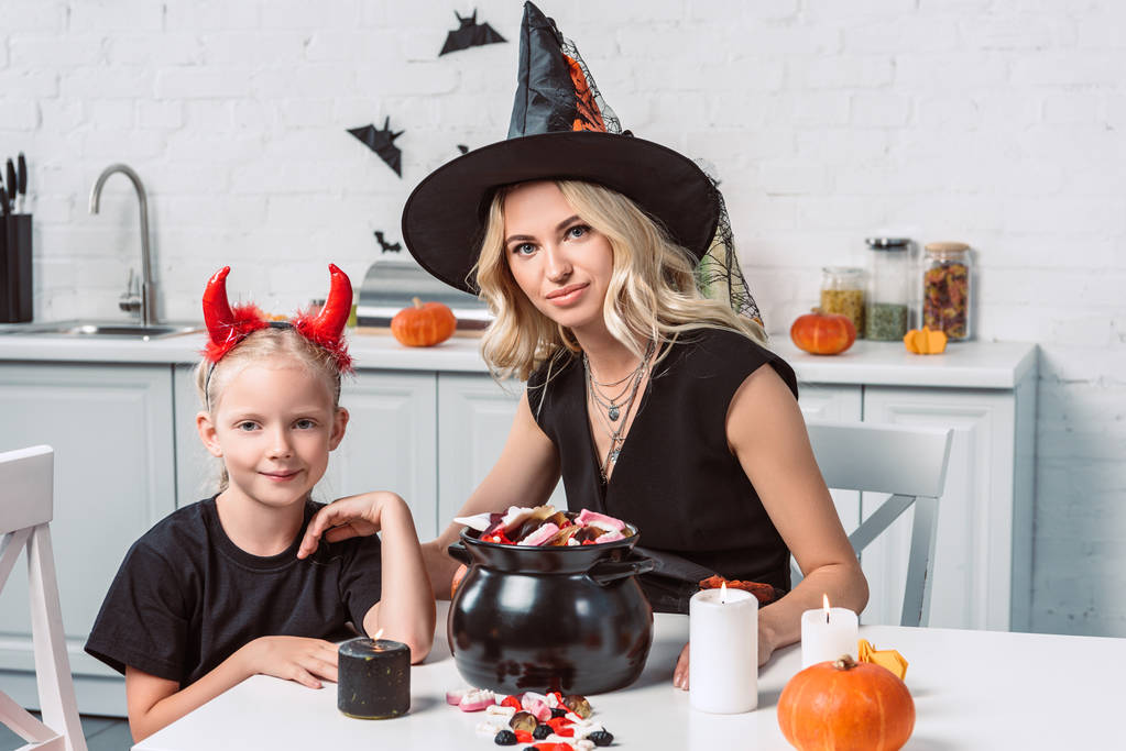 мать и маленькая дочь в костюмах на Хэллоуин за столом с угощениями в черном горшочке на кухне
 - Фото, изображение