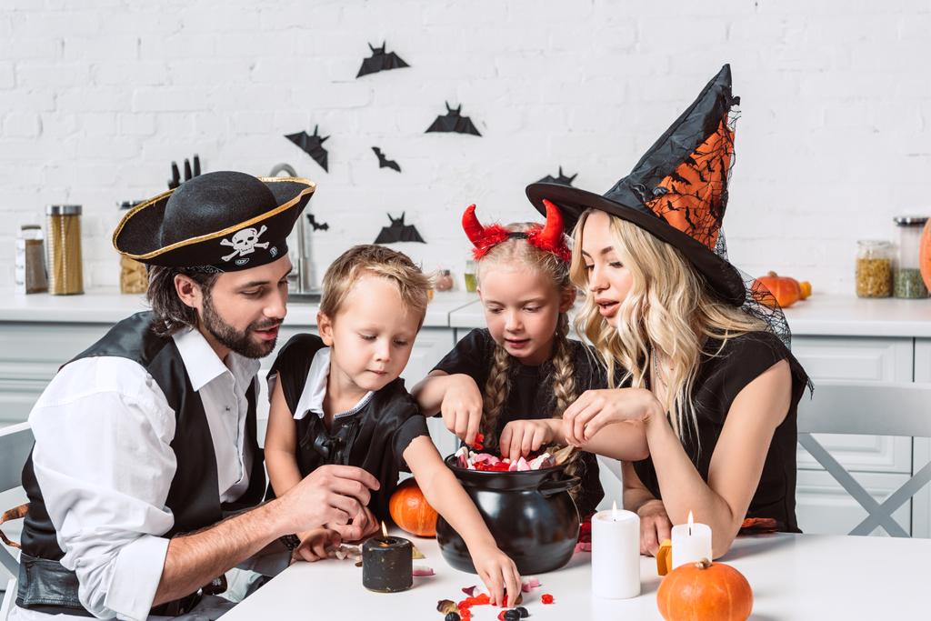 familia en varios disfraces de Halloween en la mesa con olla negra con golosinas en la cocina en casa
 - Foto, Imagen