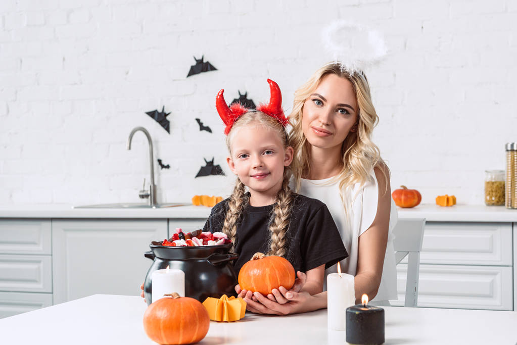 portrait of mother and daughter in halloween costumes with pumpkin at table with sweets in black pot in kitchen at home - Photo, Image