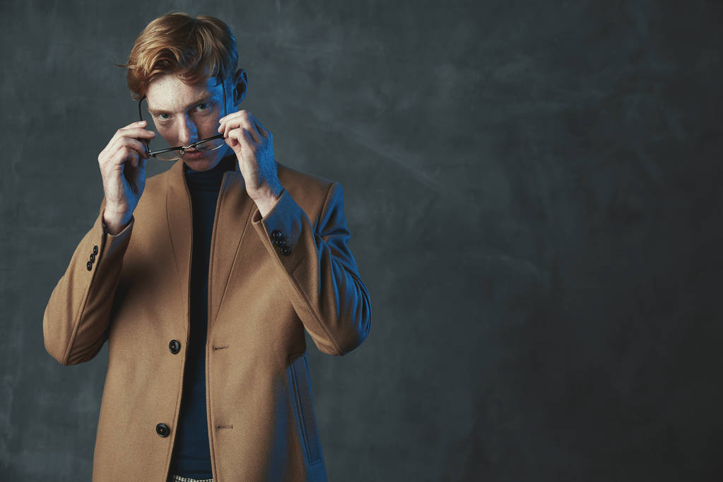 Retrato de estudio de un joven modelo masculino con pelo rojo y pecas. Vestido con abrigo marrón, con gafas. Luz azul, fondo gris
. - Foto, Imagen
