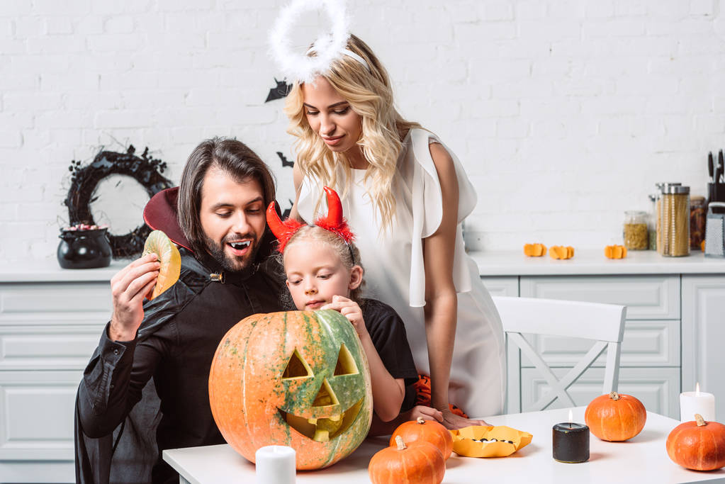 portrait of family in halloween costumes looking into pumpkin together in kitchen at home - Photo, Image