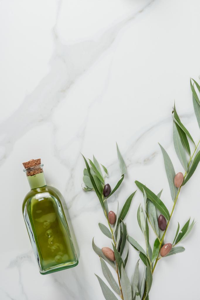 bovenaanzicht van fles zelfgemaakte olijfolie en twijgen met olijven op marmeren tafel - Foto, afbeelding