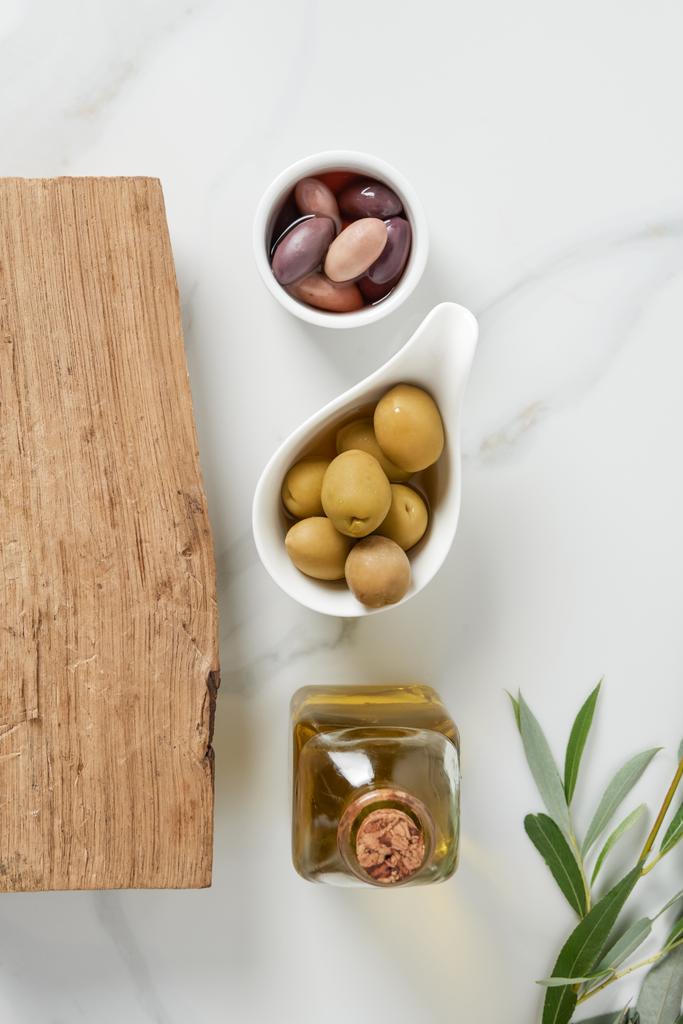bovenaanzicht van fles olijfolie, log en olijven in kommen op marmeren tafel  - Foto, afbeelding