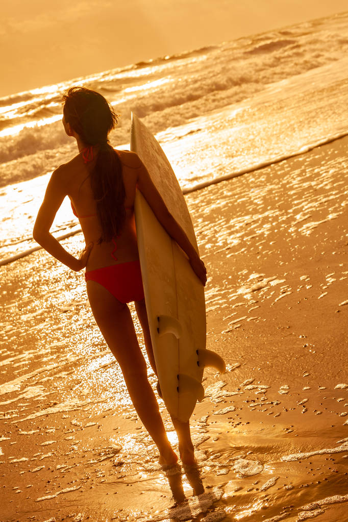 Visão traseira da bela menina surfista sexy em biquíni rosa com prancha de surf branca em uma praia ao pôr do sol ou ao nascer do sol
 - Foto, Imagem