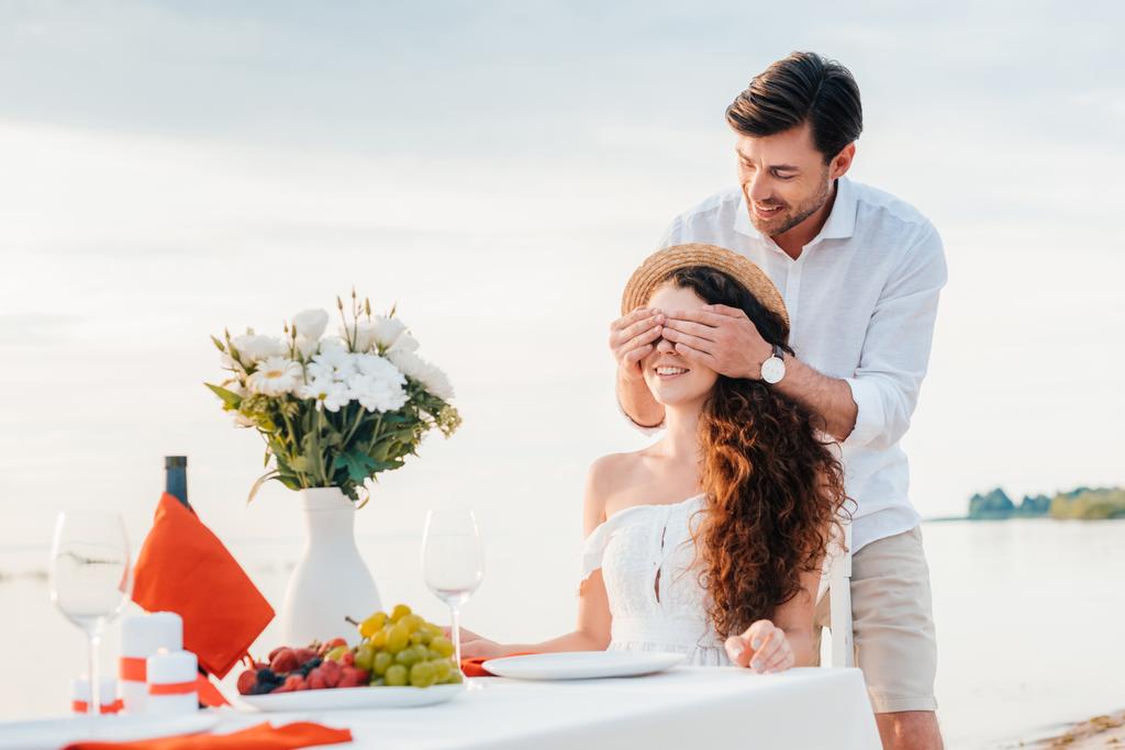 ハンサムな幸せな男は目を閉じて、ガール フレンドのための驚きを作る海岸でロマンチックなデート - 写真・画像
