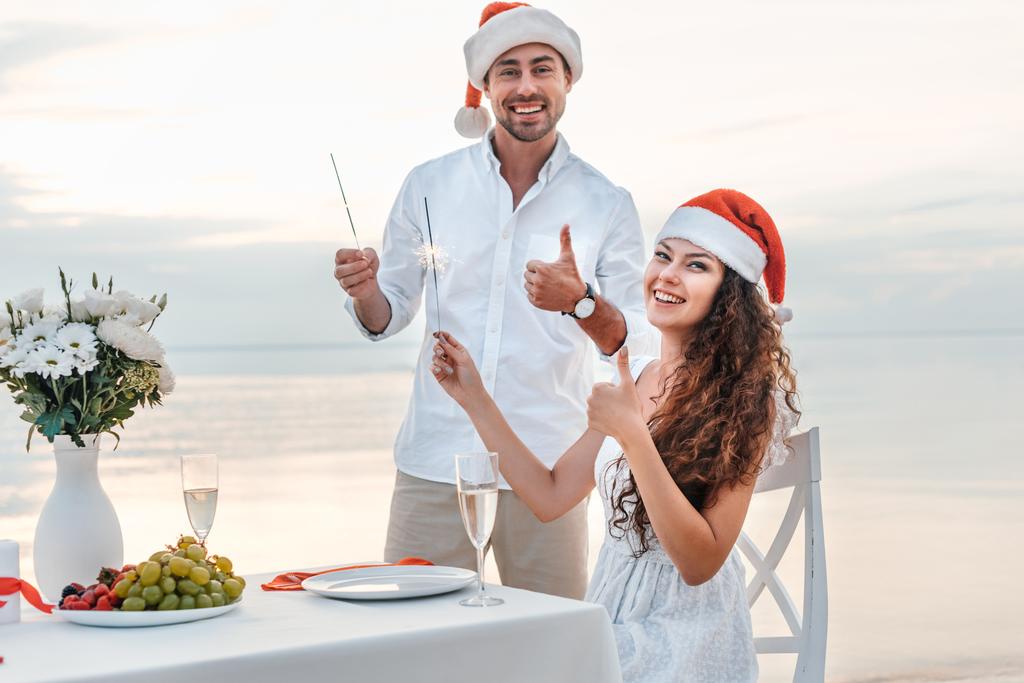 ευτυχισμένο ζευγάρι στη Σάντα καπέλα προβολή αντίχειρες επάνω και γιορτάζουμε τα Χριστούγεννα με βεγγαλικά στην παραλία - Φωτογραφία, εικόνα