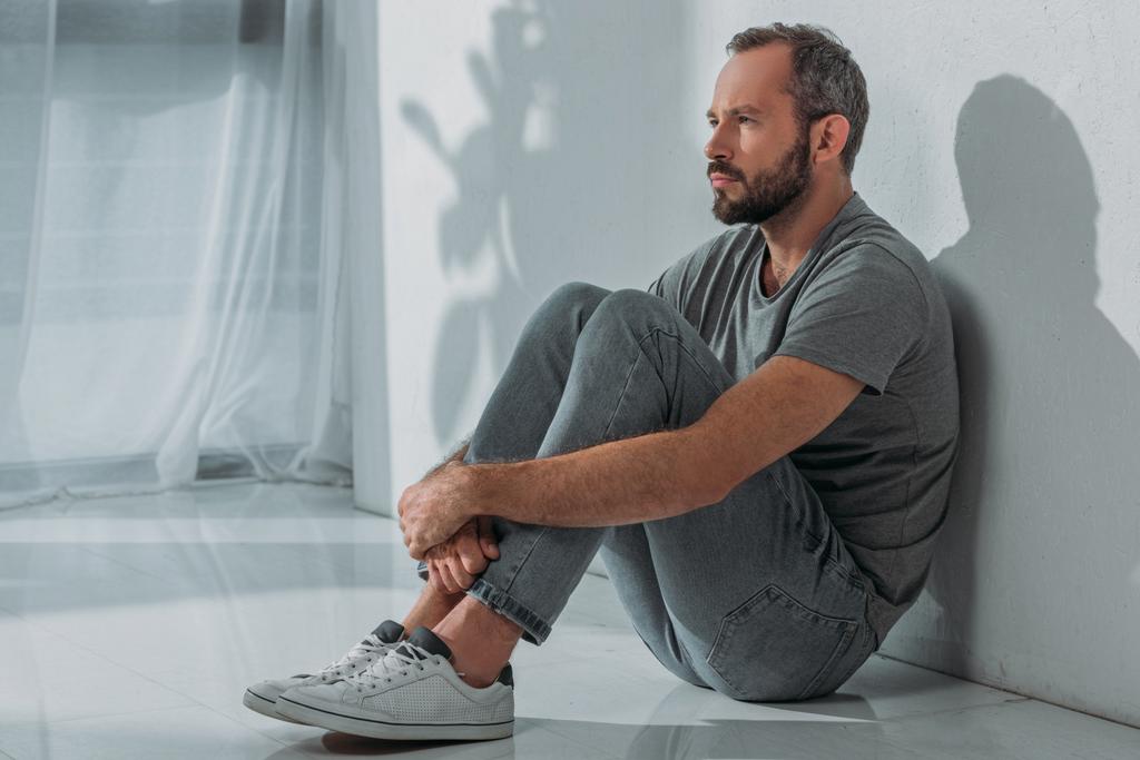 side view of sad bearded mid adult man sitting on floor and looking away - Photo, Image
