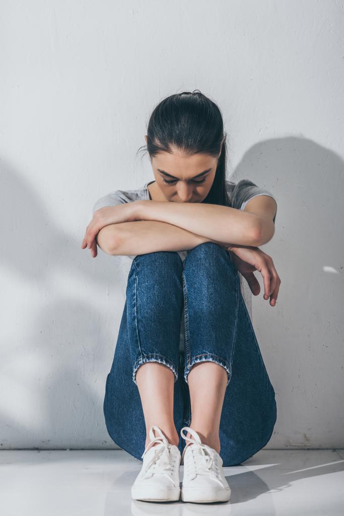 Вид в полный рост печальной стрессовой девочки, сидящей с руками на коленях на полу
 - Фото, изображение