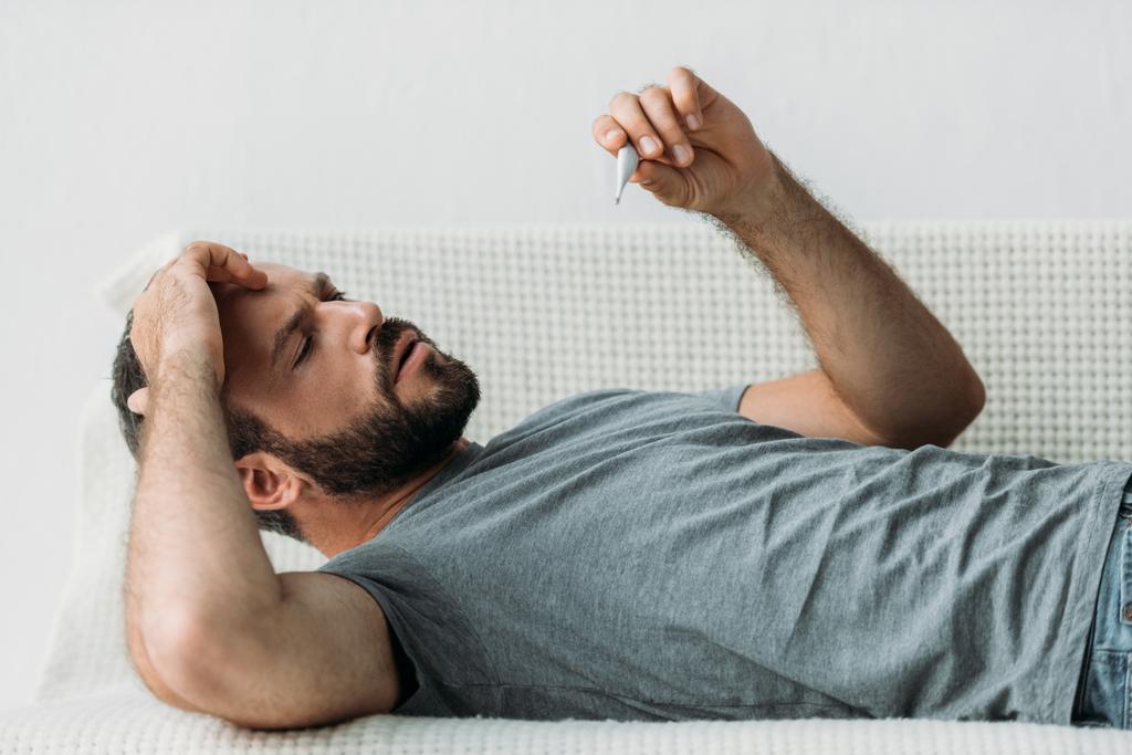 хворий бородатий чоловік лежить на дивані і тримає термометр
 - Фото, зображення