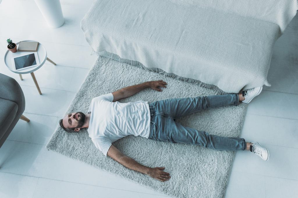 bovenaanzicht van triest bebaarde midden leeftijd man met depressie liggen op tapijt thuis - Foto, afbeelding