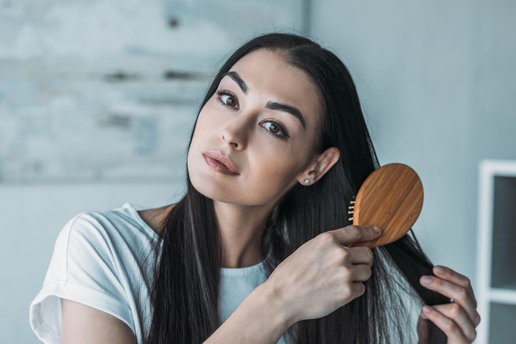 giovane donna bruna pettinando i capelli con spazzola per capelli e guardando la fotocamera
 - Foto, immagini