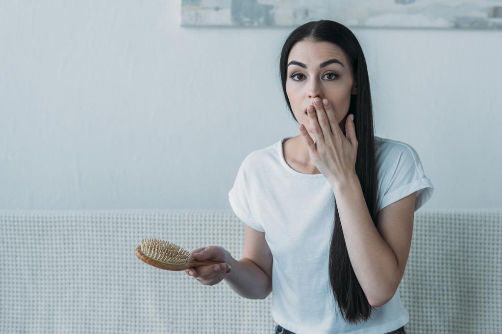 shocked brunette woman holding hairbrush and looking at camera, hair loss concept - Photo, Image