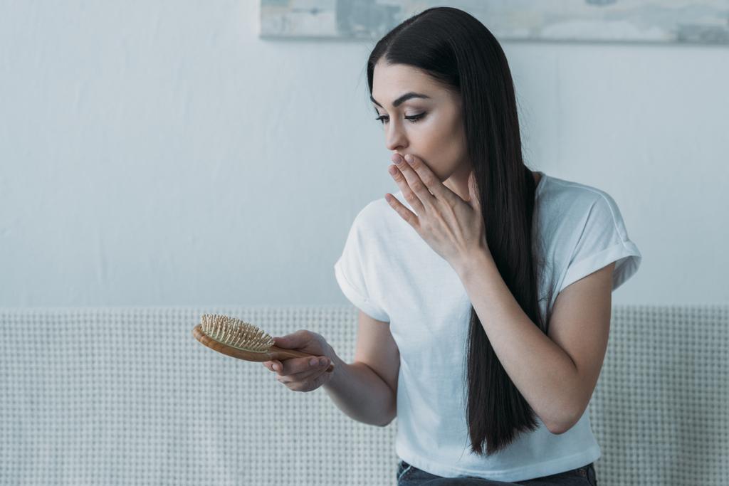 shocked brunette woman sitting on couch and looking at hairbrush, hair loss concept - Photo, Image