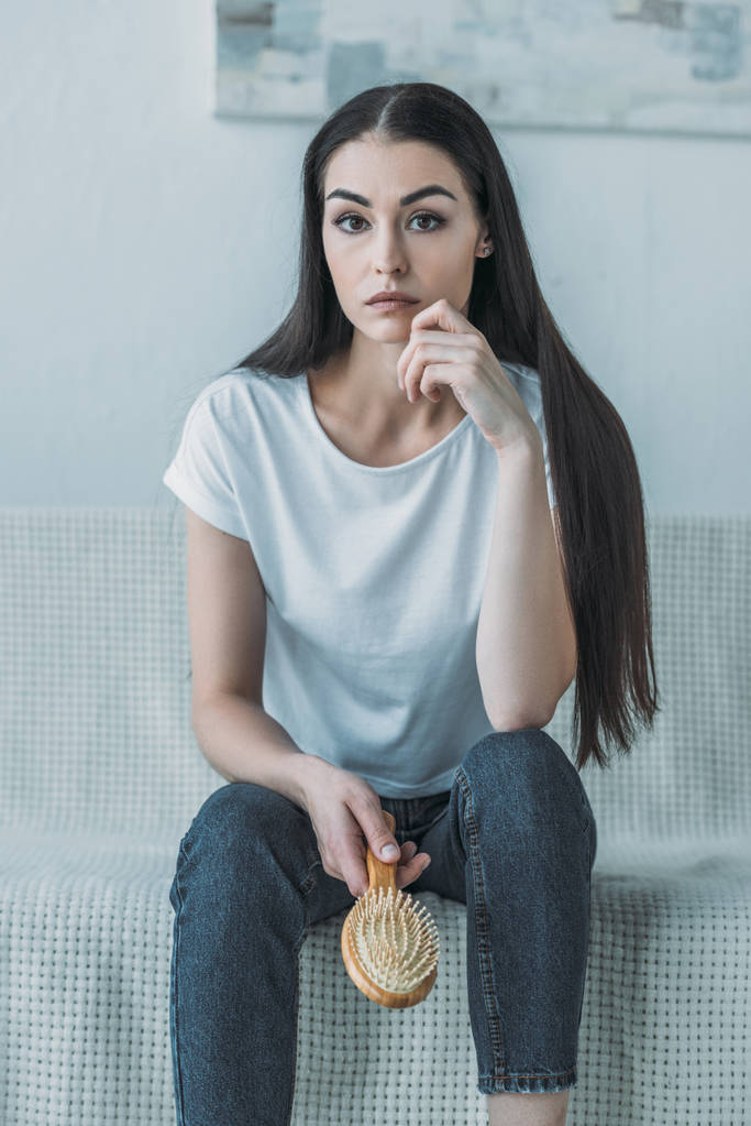 mladá bruneta žena držící kartáč na vlasy zatímco sedí na gauči a při pohledu na fotoaparát - Fotografie, Obrázek