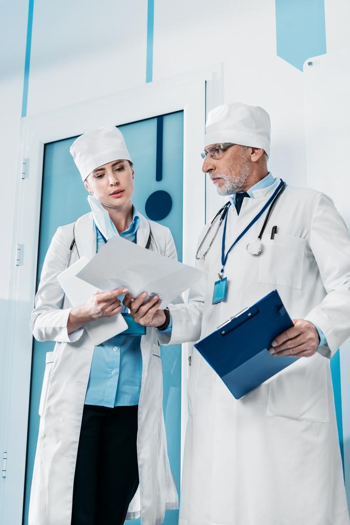 lage hoekmening van volwassen mannelijke arts tonen van papier aan vrouwelijke collega in de bandbreedte van het ziekenhuis  - Foto, afbeelding