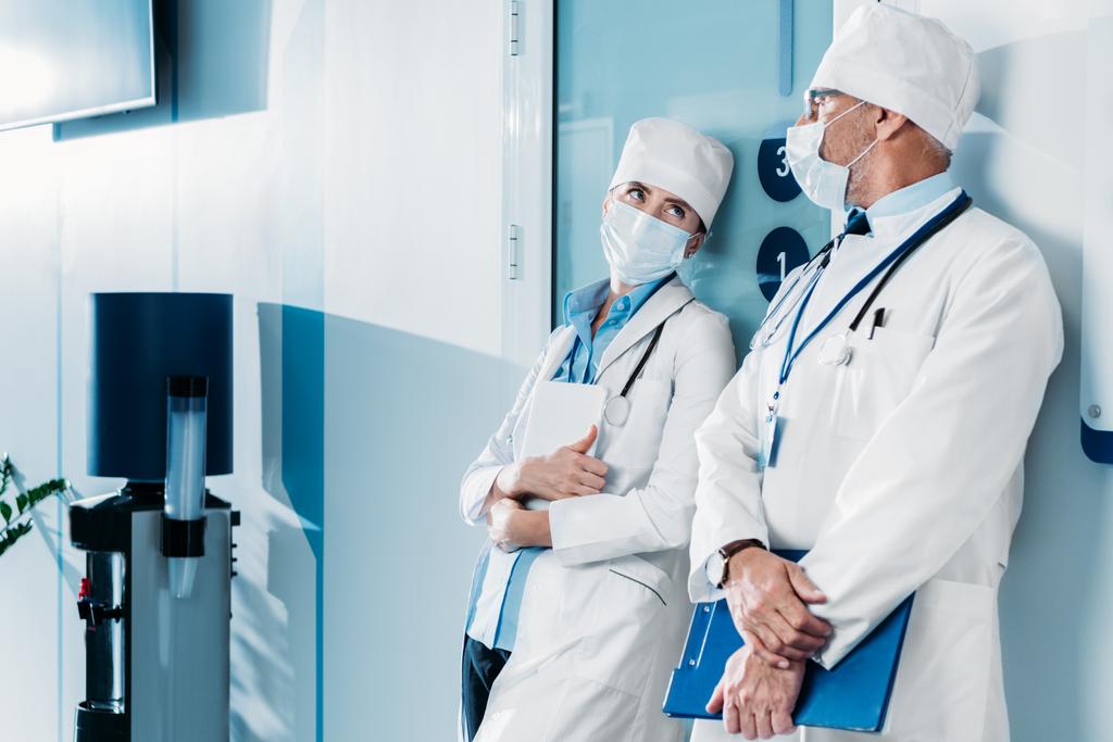 чоловічі та жіночі лікарі в медичних масках, що стоять з буфером і цифровим планшетом в лікарняному коридорі
  - Фото, зображення
