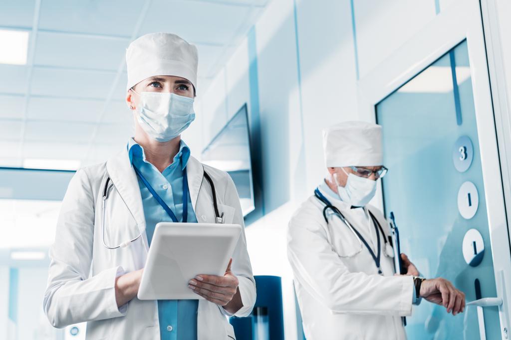 病院の廊下でクリップボードの後ろに立って彼女の男性同僚間デジタル タブレットを使用して医療のマスクで女医のローアングル ビュー  - 写真・画像