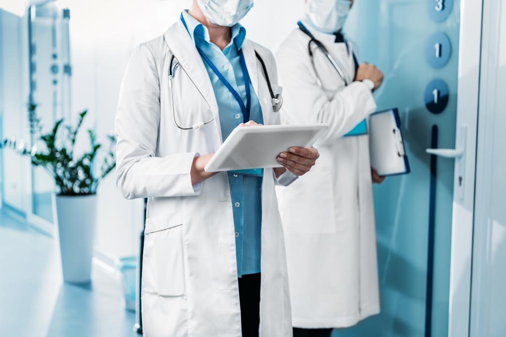 Μερική άποψη της γυναίκα γιατρό σε ιατρική μάσκα χρησιμοποιώντας ψηφιακό tablet, ενώ ο συνάδελφός της αρσενικό στέκεται πίσω από με πρόχειρο στο διάδρομο του νοσοκομείου  - Φωτογραφία, εικόνα