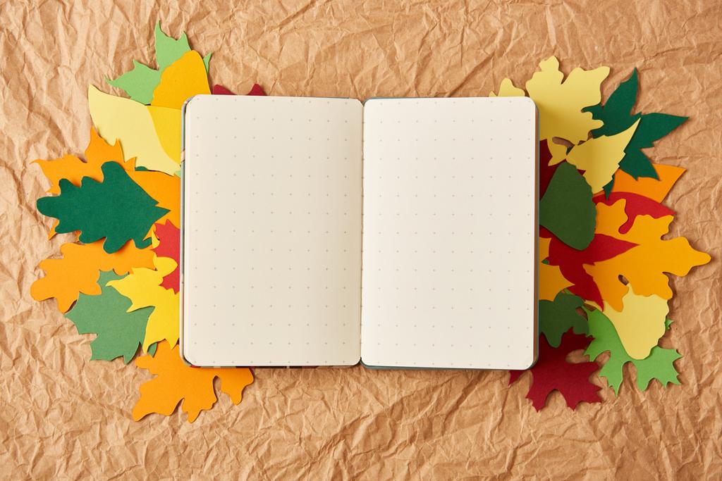 top view of empty notebook and colorful handmade paper leaves on crumpled paper backdrop - Photo, Image
