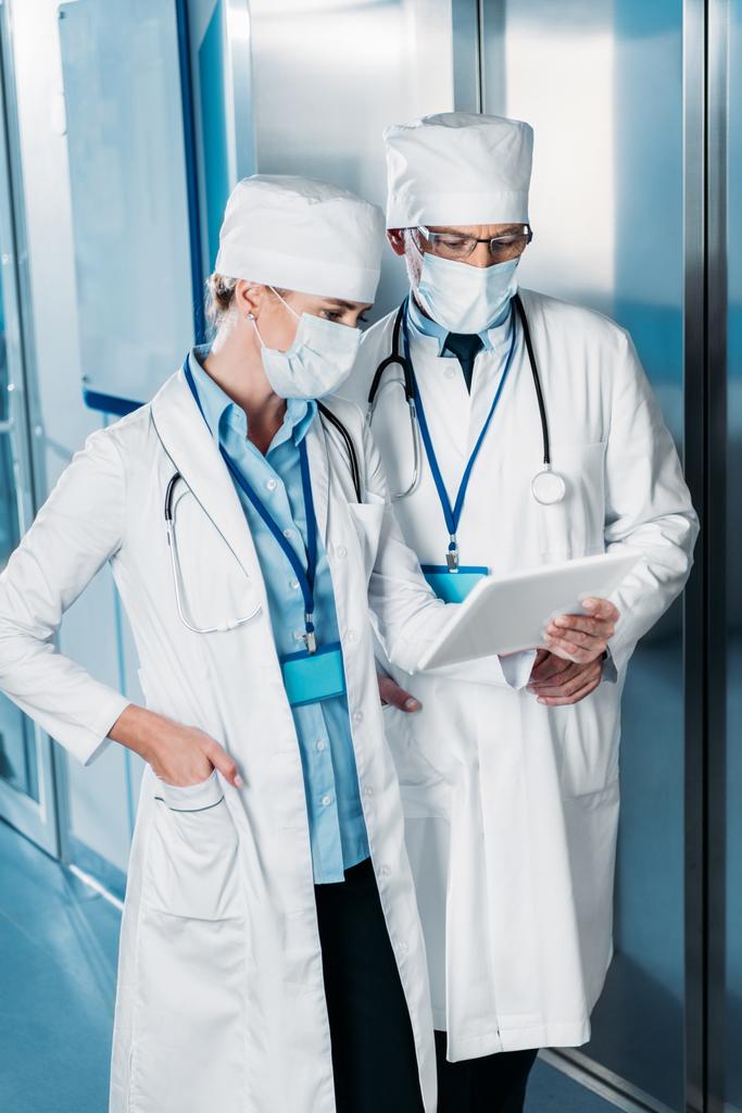 γυναίκα γιατρό σε ιατρική μάσκα δείχνει ψηφιακή δισκίο αρσενικό συνάδελφο στο διάδρομο του νοσοκομείου - Φωτογραφία, εικόνα