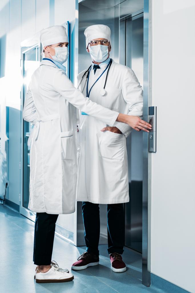 Mies- ja naislääkärit lääketieteellisissä naamioissa hissin painike sairaalan käytävällä
  - Valokuva, kuva