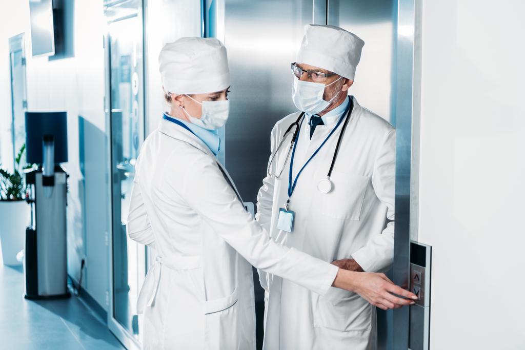 lékařka v lékařské maska mačkání tlačítka výtahu a mluví s mužského kolegu v nemocniční chodbě  - Fotografie, Obrázek
