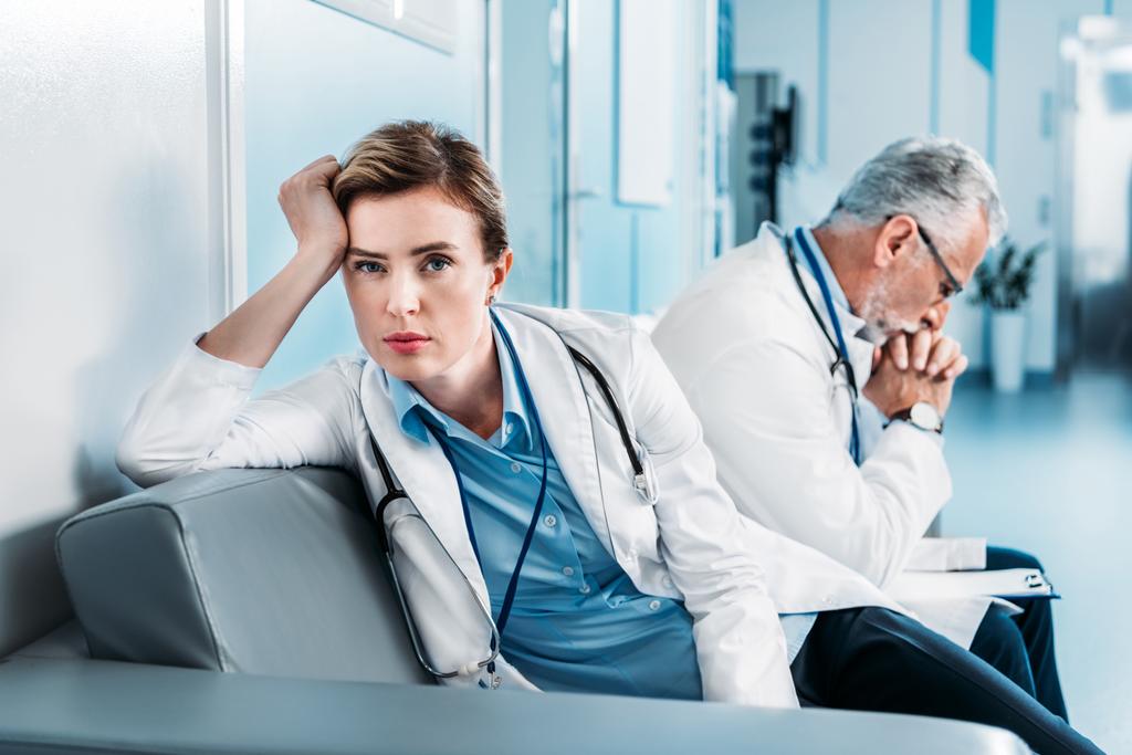 fatigué femme médecin regardant caméra sur canapé tandis que son collègue masculin assis derrière dans le couloir de l'hôpital
  - Photo, image