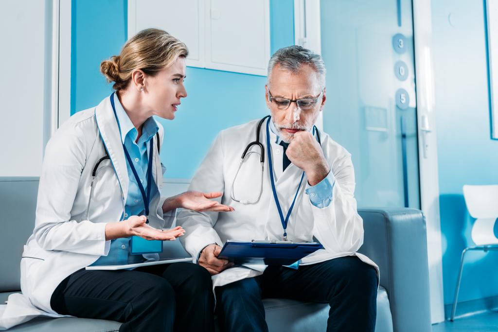 Erwachsene emotionale Ärztin gestikuliert mit den Händen und diskutiert mit männlichen Kollegen auf dem Sofa im Krankenhausflur  - Foto, Bild