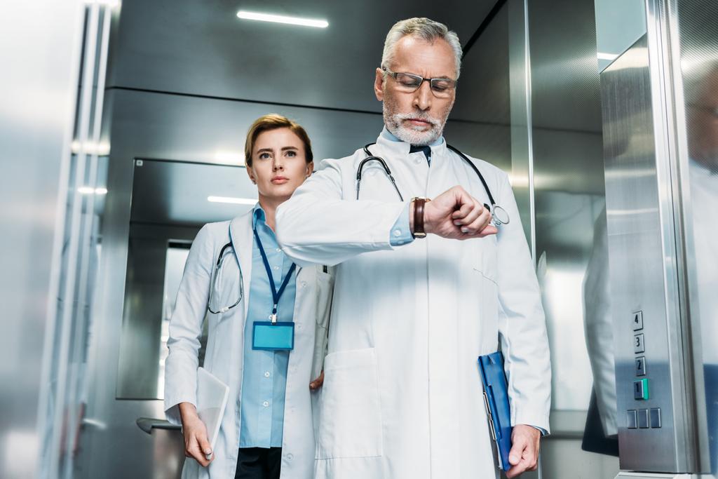 médico masculino de mediana edad serio comprobando reloj de pulsera mientras su colega mujer de pie detrás en el ascensor del hospital
  - Foto, Imagen
