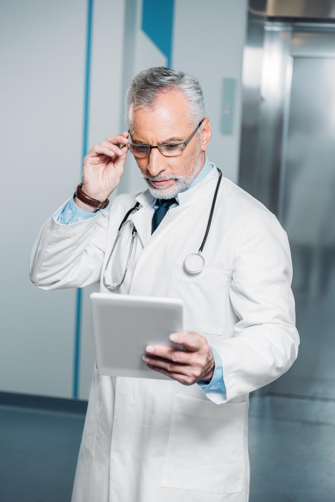 Reifer Mann mit Stethoskop über Hals, Brille und Blick auf digitales Tablet im Krankenhaus  - Foto, Bild