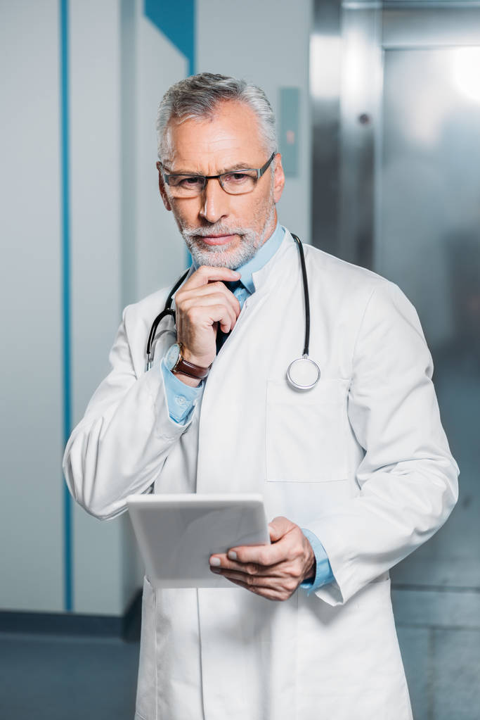 ernste reife männliche Arzt mit Stethoskop über dem Hals Blick auf Kamera und hält digitale Tablette im Krankenhaus  - Foto, Bild