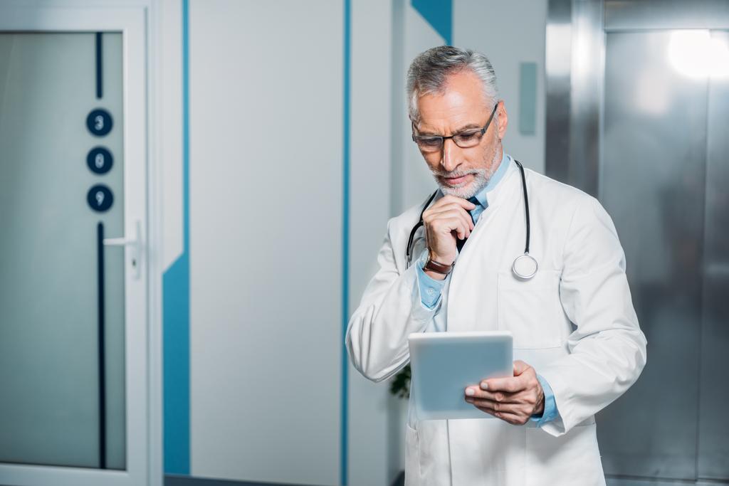 dojrzały mężczyzna lekarza z stetoskop nad szyją trzymając tablet cyfrowy w szpitalu  - Zdjęcie, obraz