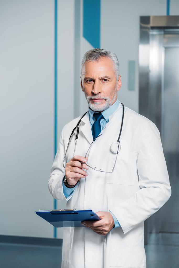 médecin masculin d'âge moyen avec stéthoscope au-dessus du cou tenant des lunettes avec presse-papiers et regardant la caméra à l'hôpital
  - Photo, image