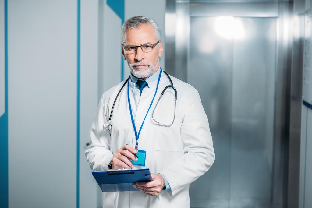 confident mature male doctor with stethoscope over neck holding clipboard and looking at camera in hospital  - Photo, Image