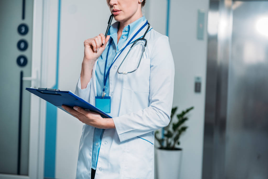 vista parcial de la doctora pensativa con portapapeles sosteniendo lápiz cerca de la boca en el hospital
  - Foto, imagen
