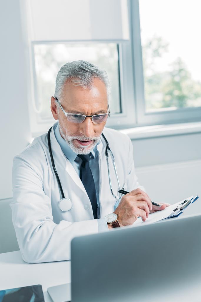 médico masculino de mediana edad escribiendo en portapapeles y mirando el portátil en la oficina
  - Foto, Imagen