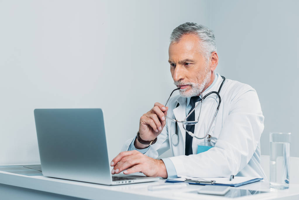 középső idős férfi orvos gazdaság szemüveg és használ laptop asztalnál hivatalban  - Fotó, kép