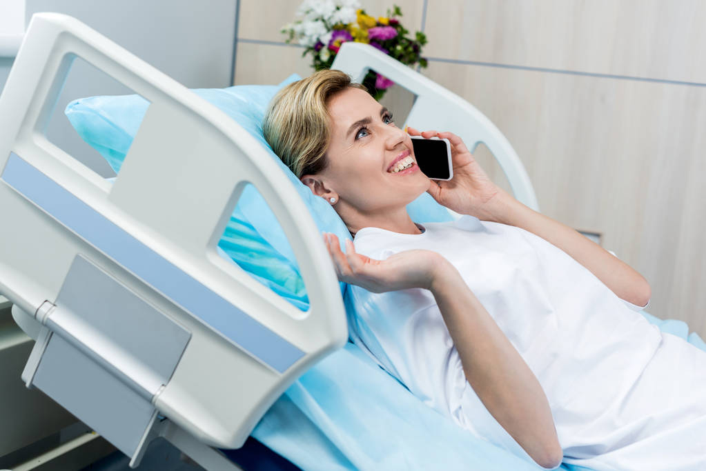 onnellinen naispotilas makaa sängyssä ja puhuu älypuhelimella sairaalahuoneessa
 - Valokuva, kuva