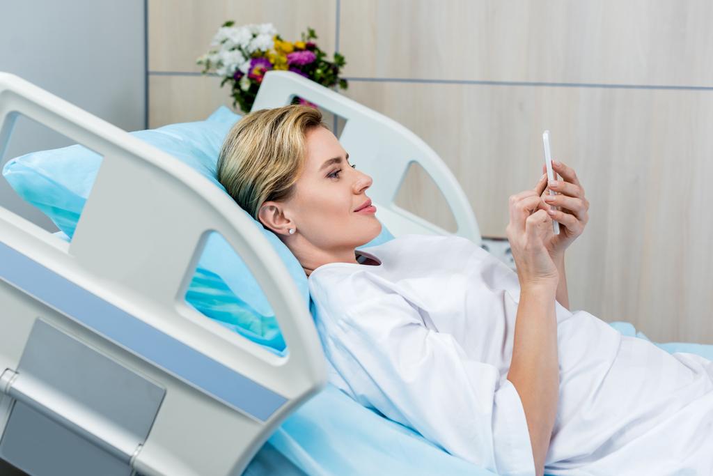 oldalnézetből a boldog felnőtt női beteg használ smartphone, a szobában kórházi ágyon  - Fotó, kép