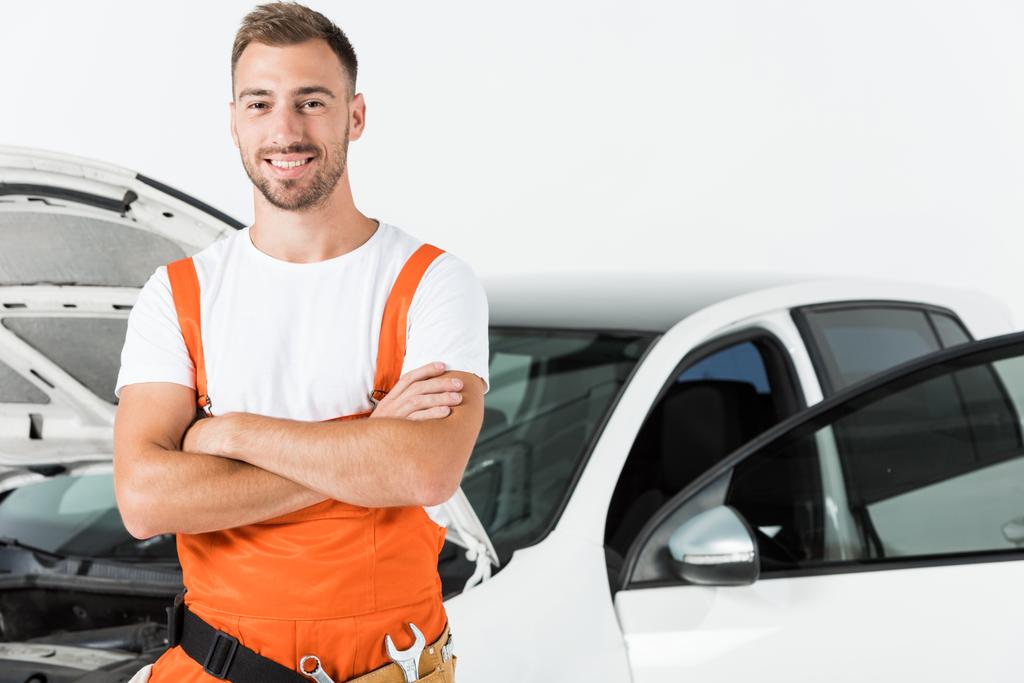 handsome auto mechanic in orange uniform standing with crossed arms near open car isolated on white - Photo, Image