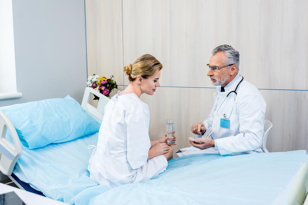 środku wieku lekarz mężczyzna z stetoskop szyję, wskazując na pigułki do pacjentki w szpitalu  - Zdjęcie, obraz
