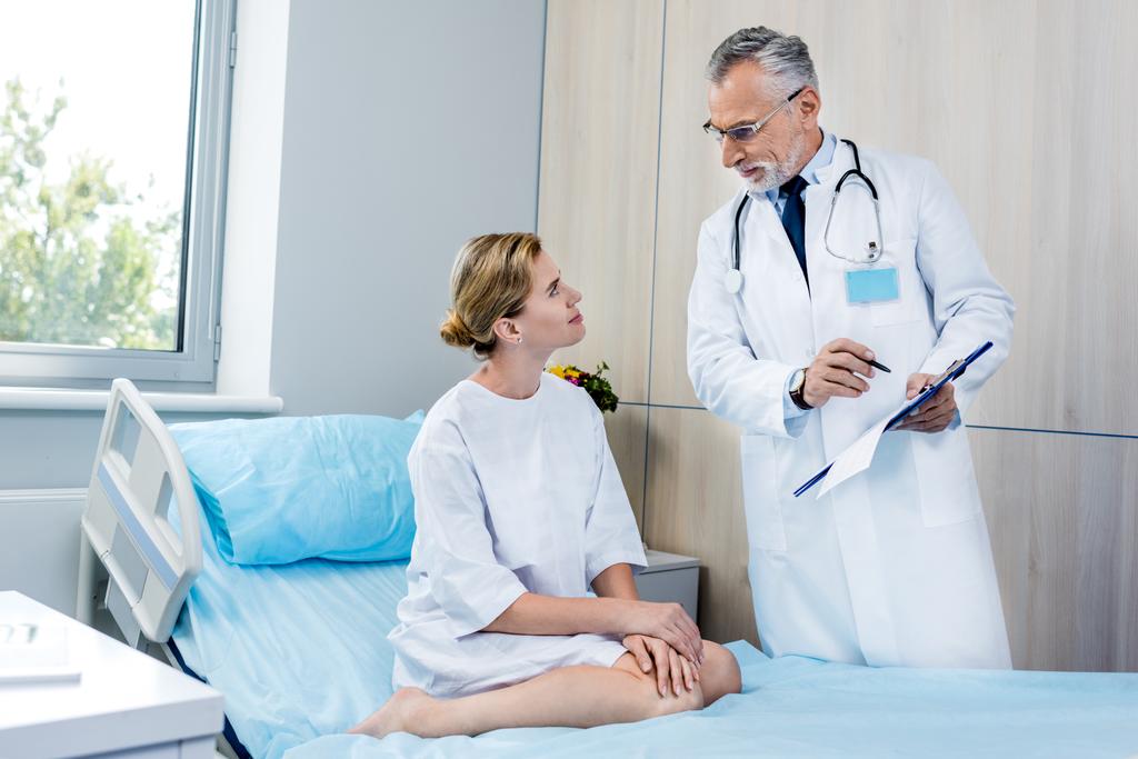 чоловічий лікар зі стетоскопом над шиєю, вказуючи в буфері до пацієнта жінки в лікарняній кімнаті
  - Фото, зображення