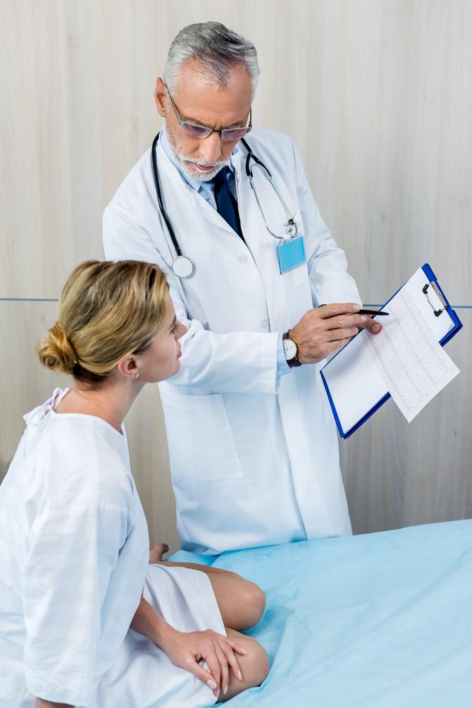 doctor masculino serio con estetoscopio sobre el cuello señalando el portapapeles al paciente femenino en la habitación del hospital
  - Foto, imagen