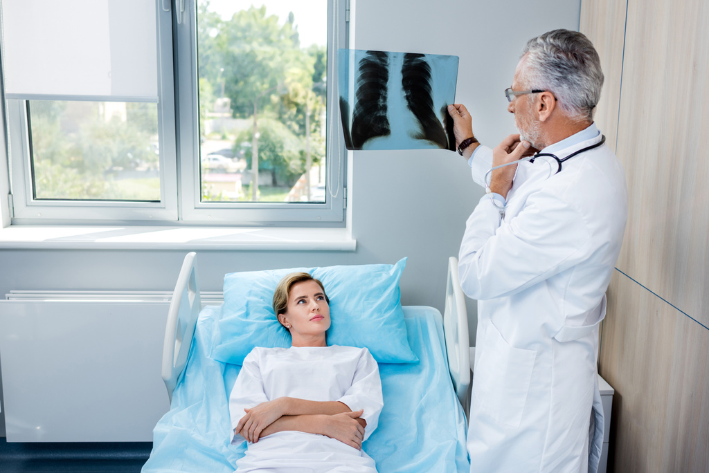 συλλογισμένος αρσενικό γιατρός, αναλύοντας εικόνα ακτίνων χ κοντά σε γυναίκα ασθενή στο δωμάτιο του νοσοκομείου - Φωτογραφία, εικόνα