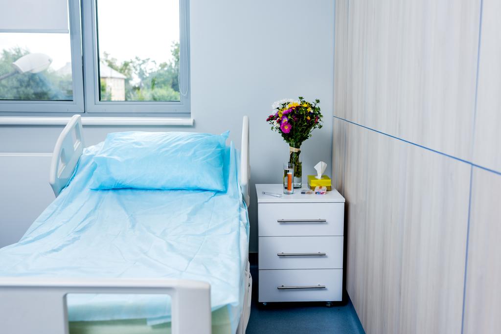Innenraum des Krankenhauszimmers mit Bett, Blumen und Nachttisch  - Foto, Bild