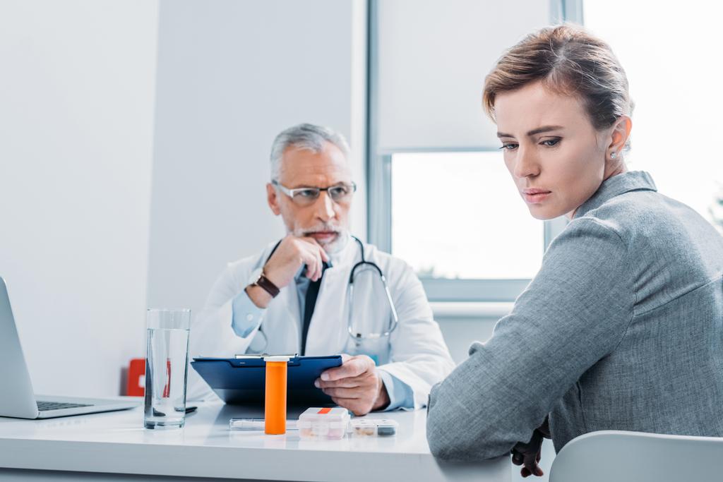 traurige Patientin schaut weg, während reifer männlicher Arzt in der Nähe am Tisch im Büro sitzt  - Foto, Bild