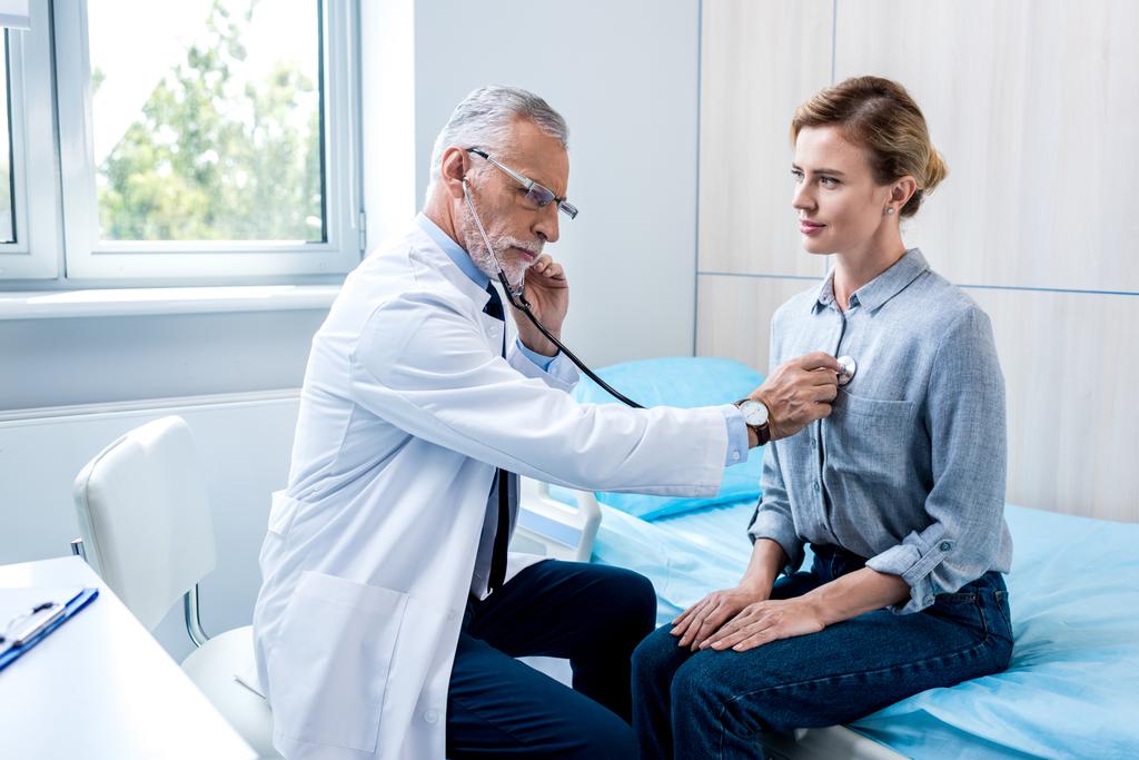 серьезный зрелый врач мужского пола осматривает пациентку стетоскопом в больничной палате
 - Фото, изображение
